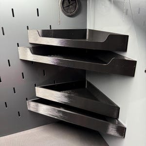 May include: Two black triangular corner shelves with raised edges, mounted on a gray pegboard wall panel. The shelves feature a textured finish and are positioned one above the other, designed to fit into a corner space for storage organization.