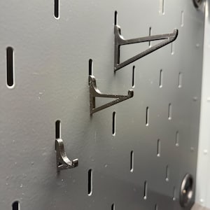 May include: A dark gray pegboard panel featuring a series of vertical slots. Three black, triangular-shaped hooks of varying lengths are inserted into the slots, extending outward from the surface to provide storage and organization for items.