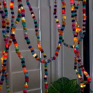 May include: Colorful beaded string lights in various colors, including red, orange, yellow, green, blue, and purple. The lights are strung together and hang from a dark metal frame. The lights are illuminated, creating a festive atmosphere.