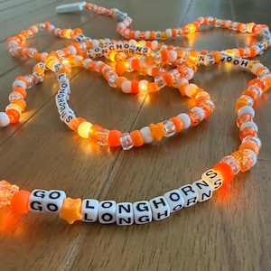 May include: A glowing orange and white beaded necklace with the words "GO LONGHORNS" and "HOOK EM" spelled out in white letter beads. The necklace has orange star-shaped beads and clear beads, and is lit with small lights.
