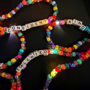 May include: Colorful beaded necklaces with illuminated beads. The necklaces feature white letter beads spelling out words like "CONFIDENT", "TALENTED", and "KIND". The beads are in various colors, including red, orange, yellow, green, blue, and purple.