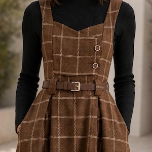 May include: A brown plaid pinafore dress featuring a square neckline, two decorative buttons on the bodice, and a matching brown belt with a gold buckle, worn over a black long-sleeved ribbed mock neck top with a fitted silhouette.