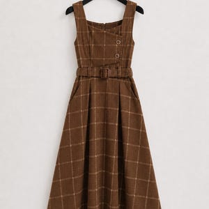 May include: A brown sleeveless midi dress featuring a white grid pattern, a square neckline, and an asymmetrical overlapping bodice detail with two circular buttons. The dress includes a matching fabric belt at the waist and a flared skirt.