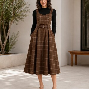May include: A brown plaid pinafore dress with a sweetheart neckline, two buttons on the bodice, and a matching belt at the waist, worn over a black long-sleeved mock neck top. The dress features a full, pleated skirt that falls below the knees.