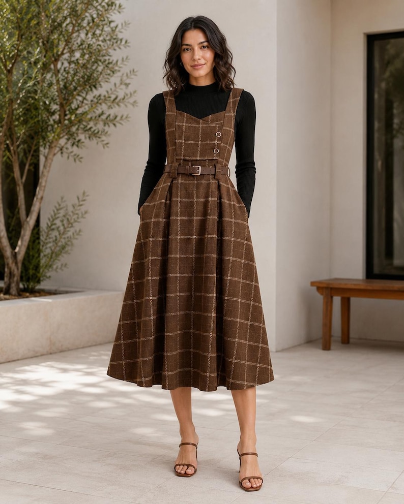 May include: A brown plaid pinafore dress with a sweetheart neckline, two buttons on the bodice, and a matching belt at the waist, worn over a black long-sleeved mock neck top. The dress features a full, pleated skirt that falls below the knees.