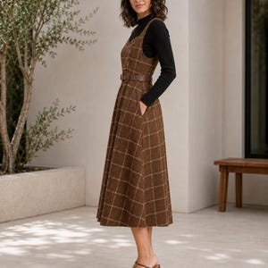 May include: A brown plaid pinafore dress featuring a V-neckline, a button-front bodice, and a matching brown belt at the waist. The dress has a full, pleated skirt with side pockets and is worn over a black long-sleeved mock neck top.