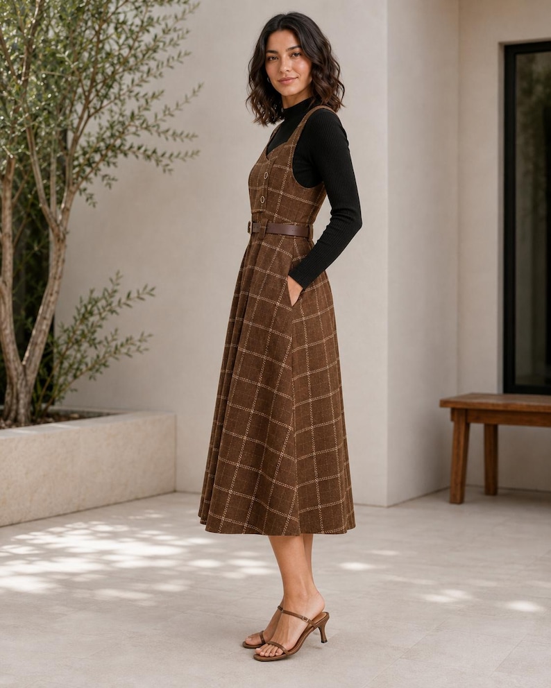 May include: A brown plaid pinafore dress featuring a V-neckline, a button-front bodice, and a matching brown belt at the waist. The dress has a full, pleated skirt with side pockets and is worn over a black long-sleeved mock neck top.