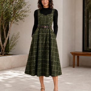 May include: A dark green plaid pinafore dress with a sweetheart neckline, two small buttons on the bodice, and a brown belt at the waist. The dress is worn over a black long-sleeved mock neck top and paired with brown open-toe heeled sandals.