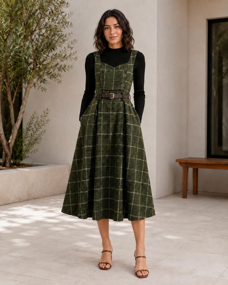 May include: A dark green plaid pinafore dress with a sweetheart neckline, two small buttons on the bodice, and a brown belt at the waist. The dress is worn over a black long-sleeved mock neck top and paired with brown open-toe heeled sandals.