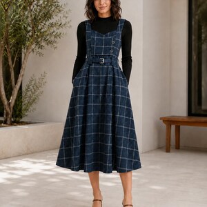 May include: A navy blue plaid pinafore dress with a sweetheart neckline, two buttons on the bodice, and a matching belt at the waist. The dress features a full, pleated skirt and is worn over a black long-sleeved mock neck top with brown sandals.