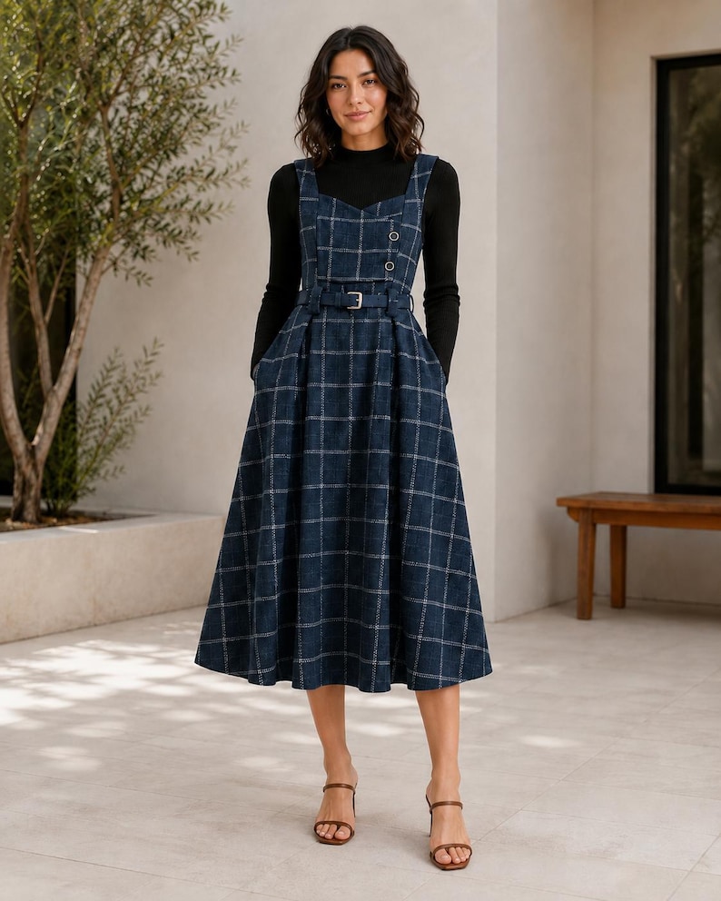 May include: A navy blue plaid pinafore dress with a sweetheart neckline, two buttons on the bodice, and a matching belt at the waist. The dress features a full, pleated skirt and is worn over a black long-sleeved mock neck top with brown sandals.