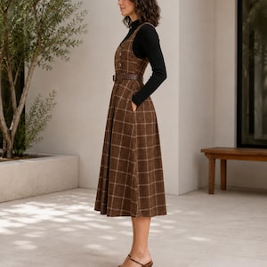 May include: A brown plaid midi-length pinafore dress featuring a button-down front, a matching brown belt at the waist, and side pockets. The dress is layered over a black long-sleeved mock neck top and paired with brown strappy heeled sandals.