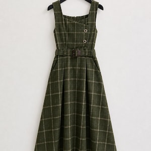 May include: A sleeveless midi dress in dark olive green with a white grid plaid pattern. The dress features a square neckline, an asymmetrical overlapping bodice with two buttons, a matching fabric belt with a buckle, and a pleated skirt.