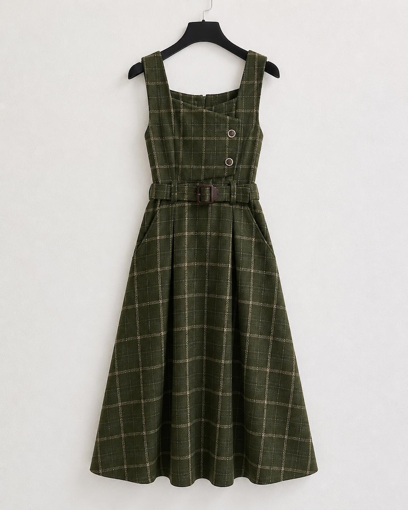 May include: A sleeveless midi dress in dark olive green with a white grid plaid pattern. The dress features a square neckline, an asymmetrical overlapping bodice with two buttons, a matching fabric belt with a buckle, and a pleated skirt.