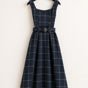 May include: A dark blue sleeveless dress featuring a white grid pattern, a square neckline, and a fitted bodice with two buttons. The dress includes a matching fabric belt with a buckle at the waist and a flared skirt that falls below the knee.