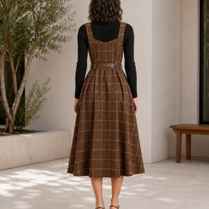 May include: A brown plaid sleeveless dress with a square neckline and a matching belt at the waist, worn over a black long-sleeved top. The dress features a pleated skirt that falls below the knee, paired with brown heeled sandals.