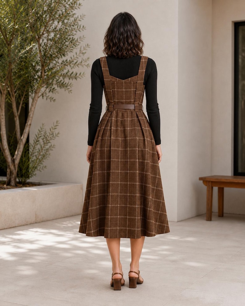 May include: A brown plaid sleeveless dress with a square neckline and a matching belt at the waist, worn over a black long-sleeved top. The dress features a pleated skirt that falls below the knee, paired with brown heeled sandals.
