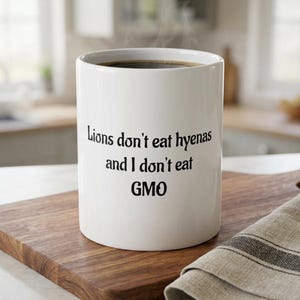 Peut inclure: Mug en céramique blanche rempli de café noir, posé sur une planche à découper en bois. Le mug porte l'inscription "Lions don't eat hyenas and I don't eat GMO" en noir. Un tissu plié à rayures grises et blanches est à côté.