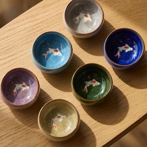 Peut inclure: Six petits bols ronds en céramique de différentes couleurs, dont bleu, vert, violet et gris. Chaque bol présente un motif de carpe koï peint au centre. Les bols sont disposés sur une surface en bois clair.