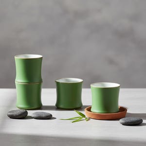 Peut inclure: Quatre tasses en céramique vertes avec un design en forme de bambou, intérieur blanc et bord doré. Une tasse est posée sur un petit dessous de verre rond marron. Les tasses sont disposées avec de petites pierres grises foncées et une feuille verte.