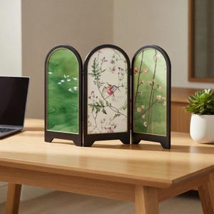 May include: A three-panel room divider with black frames and arched tops. Each panel features a different design: green with white birds, floral hummingbird, and flowering branches. The divider is set on a wooden table.