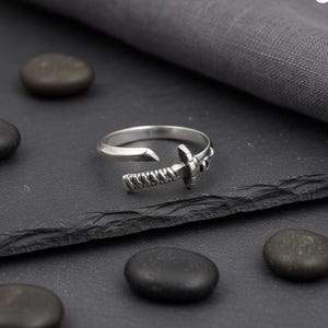 Peut inclure: Une bague argentée avec un motif d'épée miniature. La bague présente une garde et une lame détaillées, sur un fond sombre avec des pierres lisses et sombres. La bague est un bijou unique.