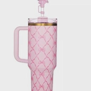 Puede incluir: Un vaso t&eacute;rmico rosa con asa y tapa en forma de lazo. El vaso presenta un patr&oacute;n de lazos repetidos en un dise&ntilde;o de celos&iacute;a. El borde tiene una banda dorada. El vaso est&aacute; dise&ntilde;ado para contener aproximadamente 1,2 litros.