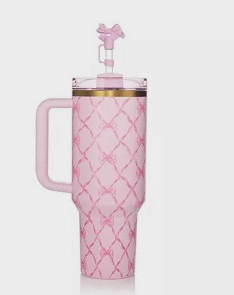 Puede incluir: Un vaso t&eacute;rmico rosa con asa y tapa en forma de lazo. El vaso presenta un patr&oacute;n de lazos repetidos en un dise&ntilde;o de celos&iacute;a. El borde tiene una banda dorada. El vaso est&aacute; dise&ntilde;ado para contener aproximadamente 1,2 litros.