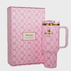 Puede incluir: Un vaso Stanley Loveshackfancy rosa con asa y caja de regalo a juego. Tanto el vaso como la caja presentan un patr&oacute;n de lazo repetido. El vaso tiene un borde dorado y el logotipo de Stanley.