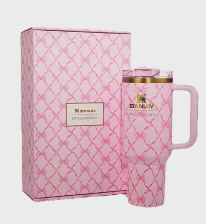 Puede incluir: Un vaso Stanley Loveshackfancy rosa con asa y caja de regalo a juego. Tanto el vaso como la caja presentan un patr&oacute;n de lazo repetido. El vaso tiene un borde dorado y el logotipo de Stanley.