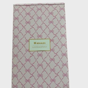 Puede incluir: Caja rectangular con un patr&oacute;n de lazo rosa y el texto "STANLEY LOVESHACKFANCY" en un rect&aacute;ngulo dorado. La caja tiene un fondo rosa claro con un patr&oacute;n repetido de lazos rosas.