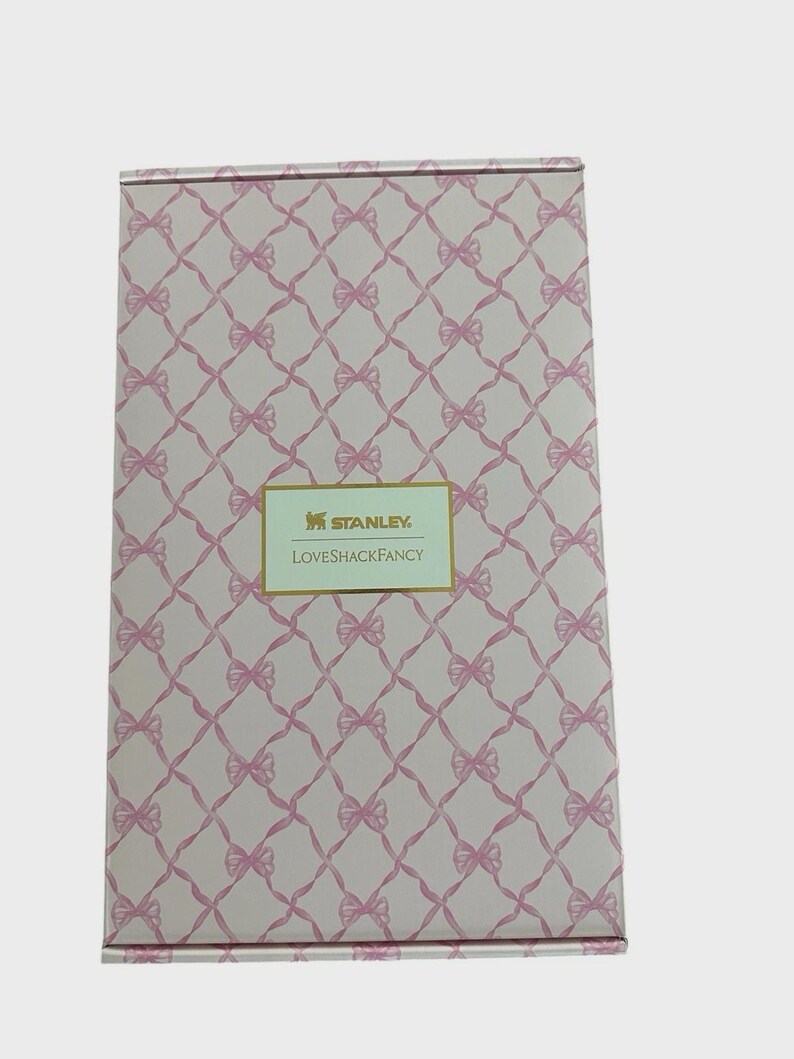 Puede incluir: Caja rectangular con un patr&oacute;n de lazo rosa y el texto "STANLEY LOVESHACKFANCY" en un rect&aacute;ngulo dorado. La caja tiene un fondo rosa claro con un patr&oacute;n repetido de lazos rosas.