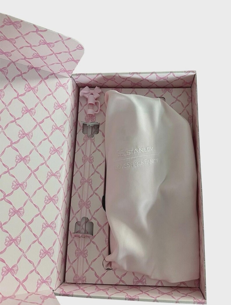 Puede incluir: Una caja de regalo rosa con un patr&oacute;n de lazo contiene una botella de agua rosa y una bolsa con cord&oacute;n a juego. El interior de la caja presenta un dise&ntilde;o de lazo repetido. La botella de agua tiene una pajita transparente y una parte superior en forma de lazo rosa. La bolsa tiene el texto "STANLEY LOVESHACKFANCY".