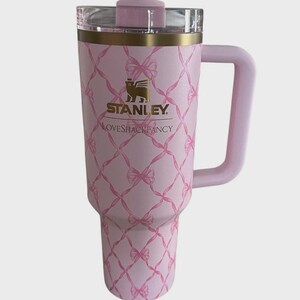 Puede incluir: Un vaso Stanley rosa con asa y tapa transparente. El vaso presenta un patr&oacute;n de lazo rosa y el logotipo "Stanley Loveshackfancy". El borde es dorado.