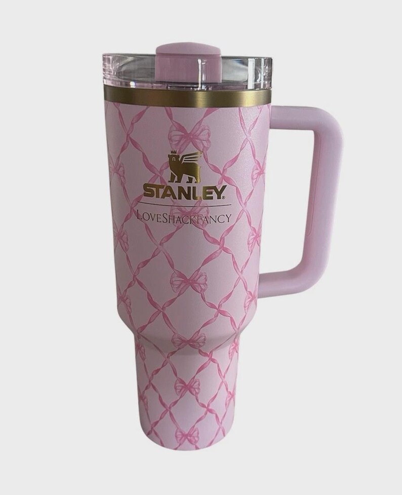 Puede incluir: Un vaso Stanley rosa con asa y tapa transparente. El vaso presenta un patr&oacute;n de lazo rosa y el logotipo "Stanley Loveshackfancy". El borde es dorado.