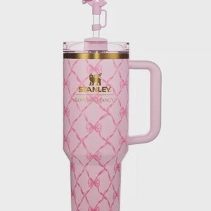 Puede incluir: Un vaso Stanley rosa con asa y un adorno en forma de lazo. El vaso presenta un patr&oacute;n de diamantes con lazos rosas y detalles dorados. El texto "STANLEY LOVESHACKFANCY" est&aacute; impreso en la parte delantera.