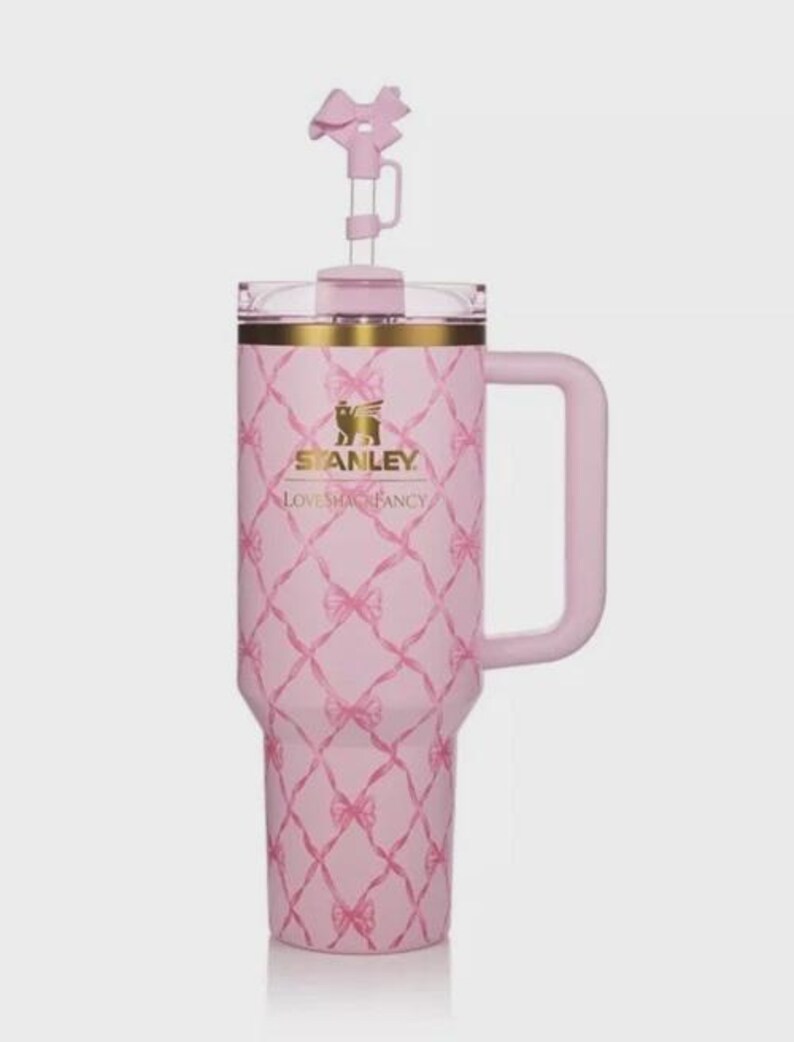 Puede incluir: Un vaso Stanley rosa con asa y un adorno en forma de lazo. El vaso presenta un patr&oacute;n de diamantes con lazos rosas y detalles dorados. El texto "STANLEY LOVESHACKFANCY" est&aacute; impreso en la parte delantera.