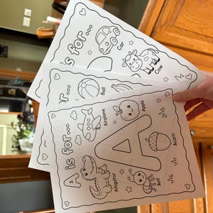May include: A stack of three white coloring pages featuring the alphabet. The top page shows a car and a cow, the middle page has a ball and an airplane, and the bottom page displays the letter "A" with an alligator, apple, and ant.