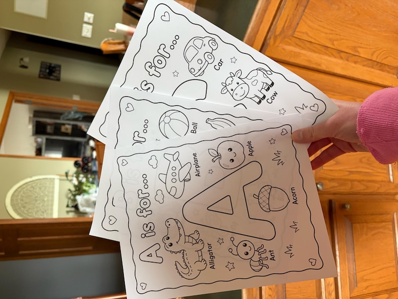 May include: A stack of three white coloring pages featuring the alphabet. The top page shows a car and a cow, the middle page has a ball and an airplane, and the bottom page displays the letter "A" with an alligator, apple, and ant.