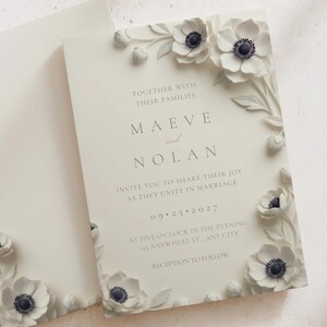 Op de afbeelding: Ivoor trouwkaart met reliëf witte anemoonbloemen en tekst. De uitnodiging luidt "Maeve en Nolan" en bevat de trouwdatum en -tijd. Een bijpassende envelop is zichtbaar.
