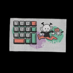 Puede incluir: Un parche rectangular de tela tejida con fondo gris claro. Presenta un gráfico de teclado numérico negro, un personaje de dibujos animados y un gato pequeño. El texto PLANETE NET está impreso en la parte inferior, con texto adicional visible.