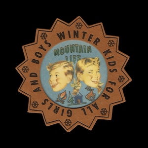 Peut inclure: Un écusson en cuir beige en forme d'étoile avec un centre circulaire bleu clair portant le texte MOUNTAIN LIFE. L'écusson présente des illustrations de deux enfants, un feu de camp, un chalet et un cerf. Le texte en bordure indique WINTER KIDS FOR ALL GIRLS AND BOYS.