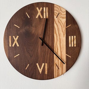 Könnte beinhalten: Eine runde Wanduhr aus Holz mit zweifarbigem Design. Das Zifferblatt hat römische Ziffern und Minutenmarkierungen. Die Zeiger der Uhr sind schwarz. Das Holz hat eine natürliche Maserung und eine polierte Oberfläche.