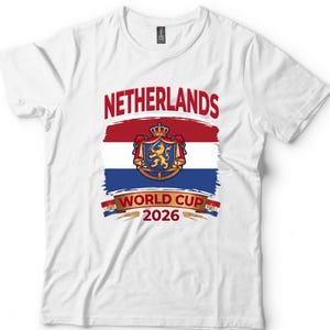 May include: White t-shirt featuring a design for the Netherlands World Cup 2026. The design includes the word "NETHERLANDS" in red, a crest, and the text "WORLD CUP 2026". The shirt has a crew neck and short sleeves.