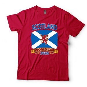 May include: A red t-shirt featuring the word "SCOTLAND" above a blue and white flag design with a red lion emblem. Below the flag is the text "WORLD CUP 2026". The shirt is made of a soft material.