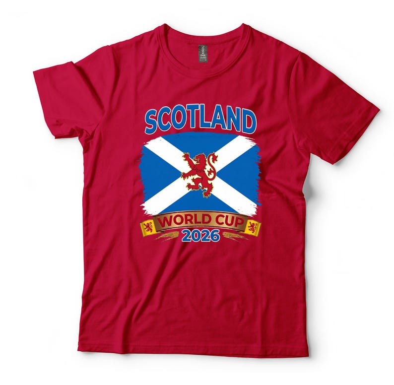 May include: A red t-shirt featuring the word "SCOTLAND" above a blue and white flag design with a red lion emblem. Below the flag is the text "WORLD CUP 2026". The shirt is made of a soft material.