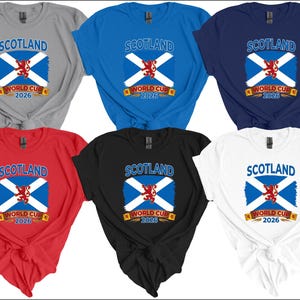 May include: Six t-shirts in various colors: gray, blue, navy, red, black, and white. Each shirt features the word "SCOTLAND" above a design of the Scottish flag and the text "WORLD CUP 2026".