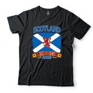 May include: Black t-shirt featuring the Scottish flag with a white cross on a blue background, a red lion, and the text "SCOTLAND WORLD CUP 2026". The design is printed on the front of the shirt.