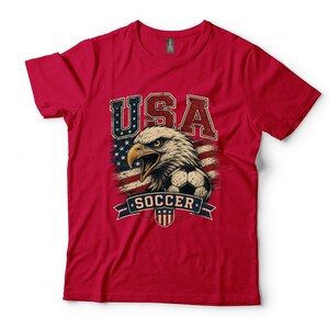 Puede incluir: Una camiseta roja de manga corta con un estampado gr&aacute;fico de una cabeza de &aacute;guila calva, un bal&oacute;n de f&uacute;tbol y un fondo de bandera estadounidense desgastada. El texto USA aparece en letras grandes sobre el &aacute;guila y la palabra SOCCER debajo.