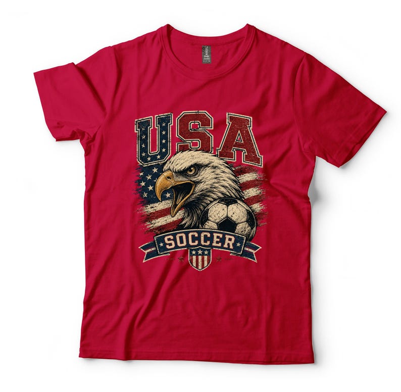 Puede incluir: Una camiseta roja de manga corta con un estampado gr&aacute;fico de una cabeza de &aacute;guila calva, un bal&oacute;n de f&uacute;tbol y un fondo de bandera estadounidense desgastada. El texto USA aparece en letras grandes sobre el &aacute;guila y la palabra SOCCER debajo.